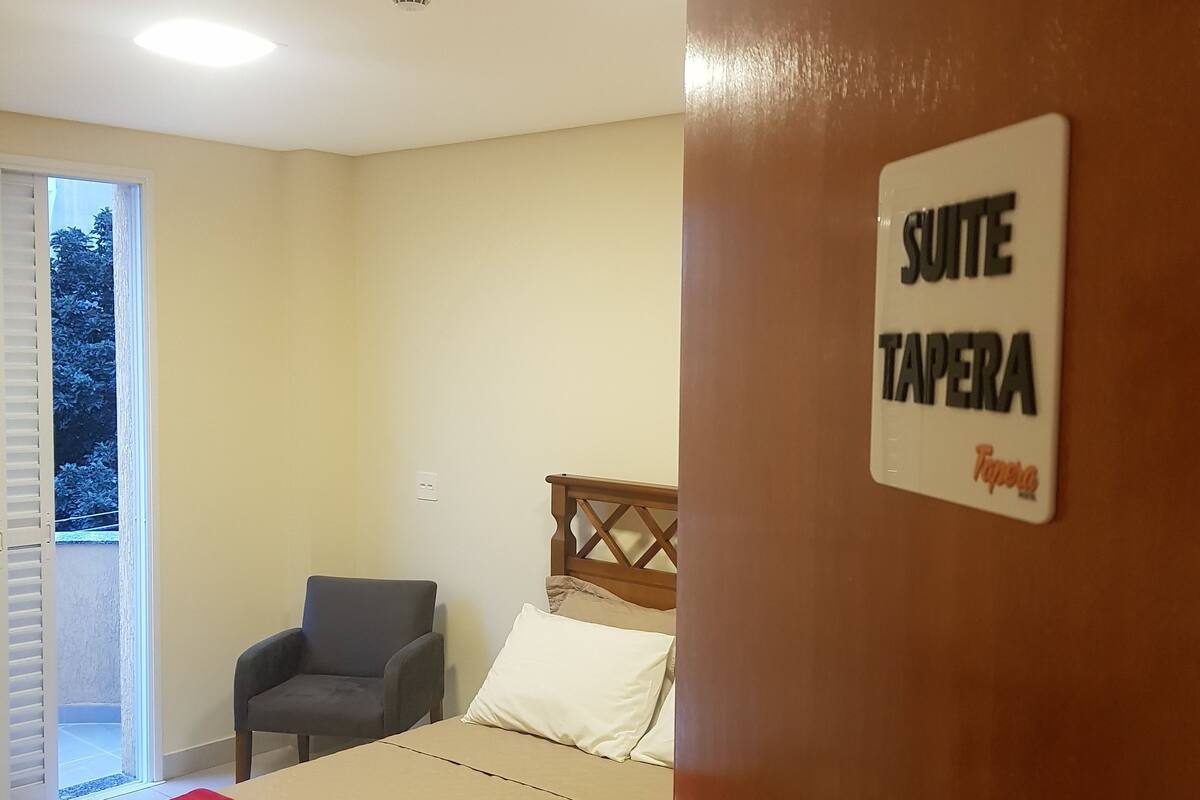 A hotel room with a bed, a chair, and a sign indicating it's a 'SUITE TAPERA'.