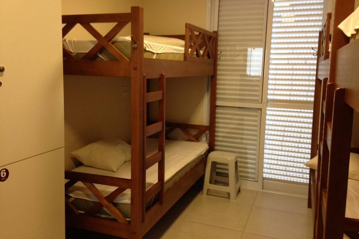 A bunk bed room with a small stool, a door, and blinds on the window.