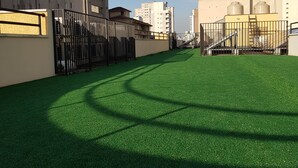 A rooftop with artificial green turf, metal fencing, and buildings in the background. - Tapera Hostel (São Paulo)