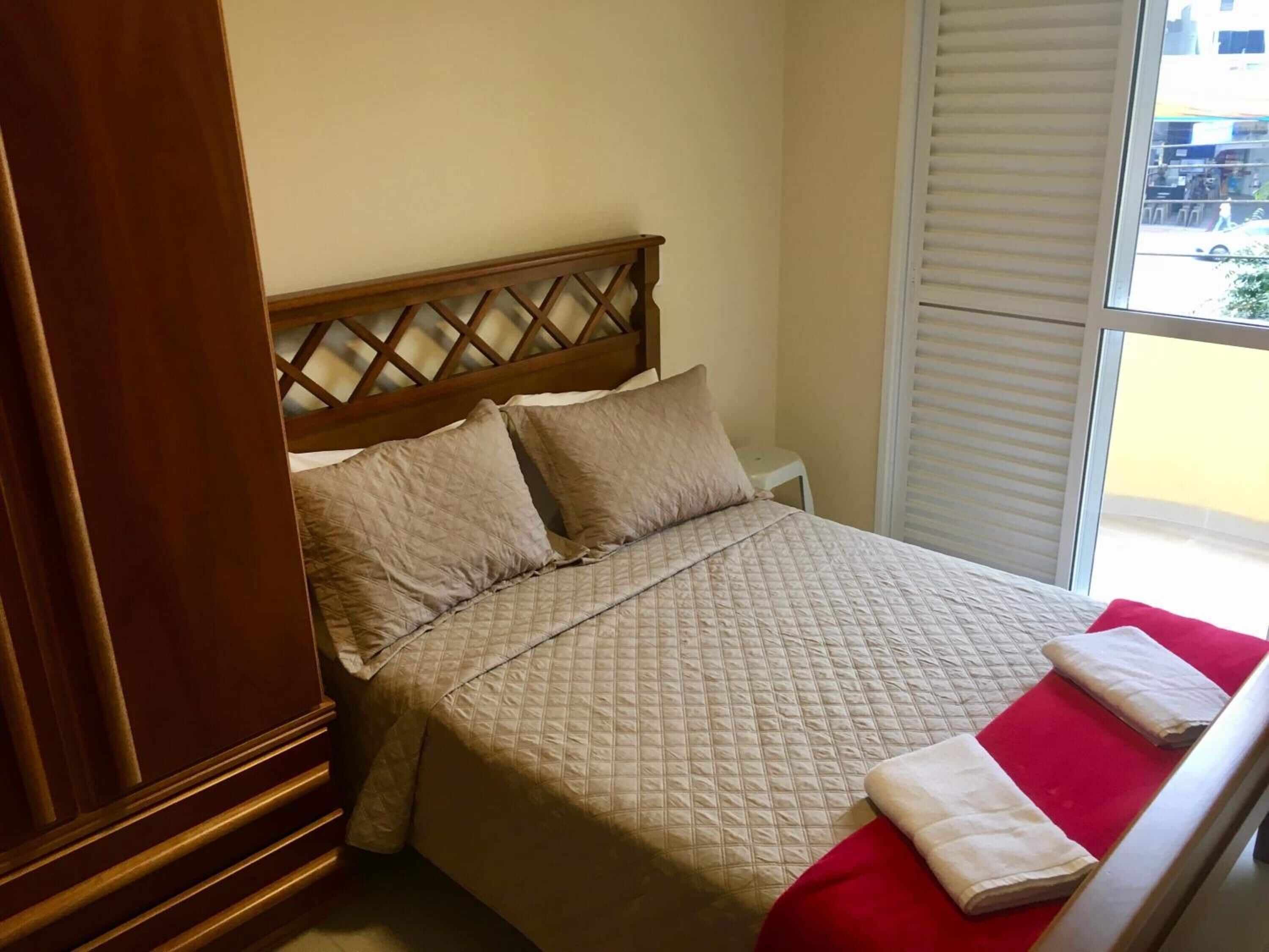 A neatly made bed with a quilted bedspread and two pillows, a wooden headboard, a small bedside table, and a window with blinds.