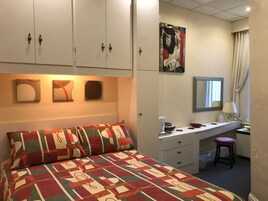 Een slaapkamer met een bed, een bureau en een stoel. Er zijn kasten boven het bed en een schilderij aan de muur.