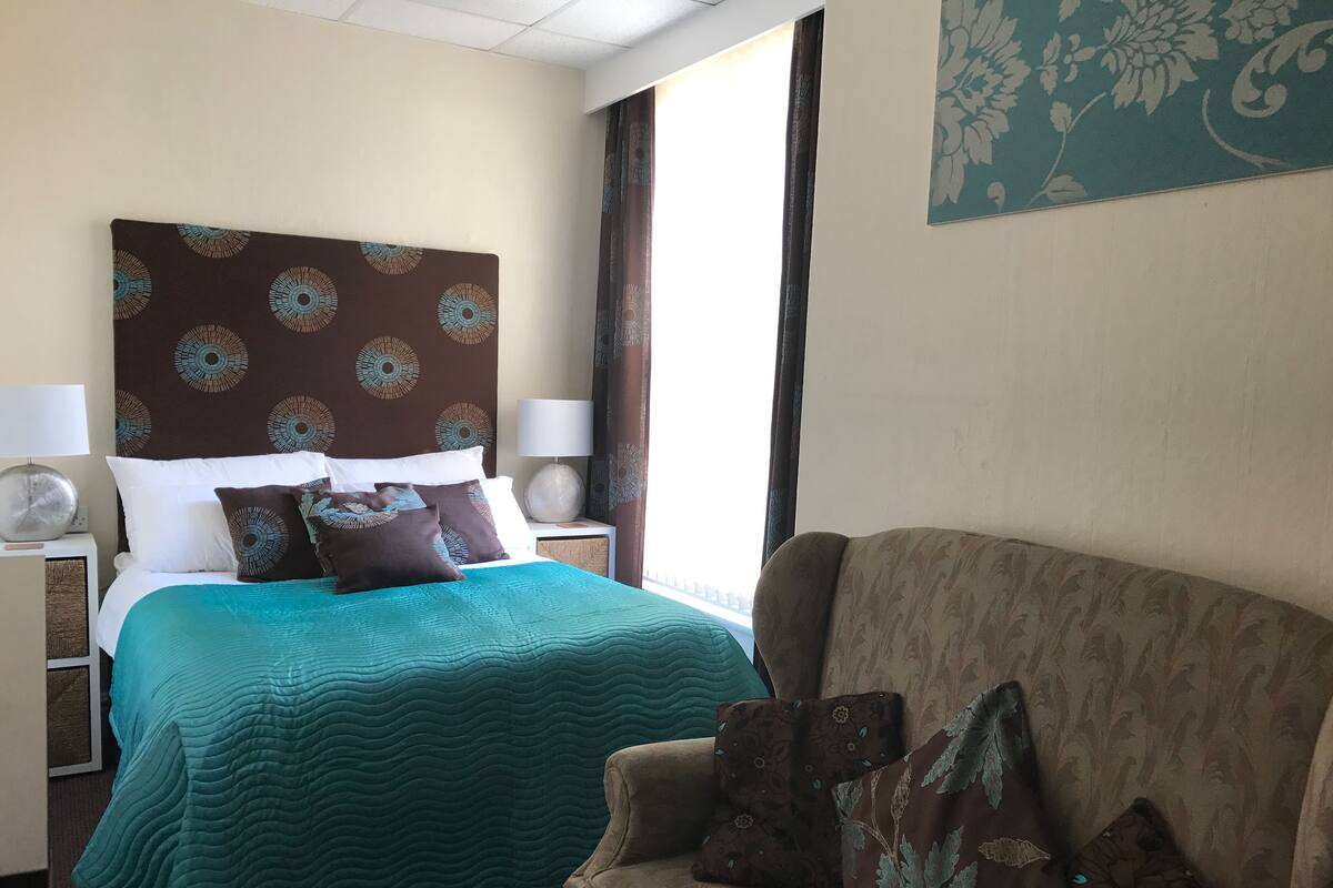 Une chambre d’hôtel avec un lit, un canapé, une table de chevet avec une lampe et une fenêtre avec des rideaux.