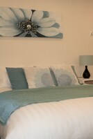 Een slaapkamer met een bed, kussens, een vaas en een schilderij aan de muur.