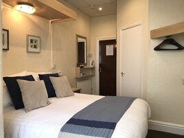 Een net opgemaakt bed met een gestreept dekbed, een nachtkastje met een lamp, een spiegel en een deur met een kapstok.