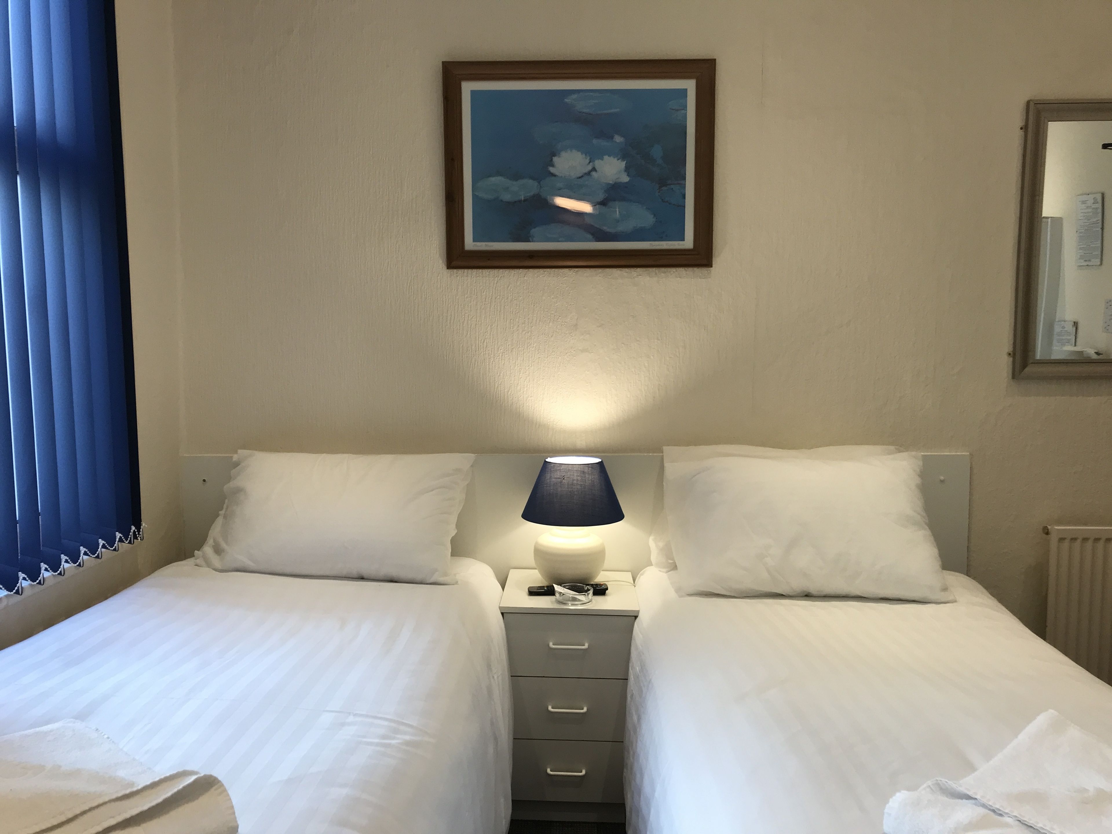 Hotellihuone, jossa on kaksi erillistä sänkyä, yöpöytä lampun kanssa, seinällä oleva kehystetty kuva ja ikkuna, jossa on siniset verhot.