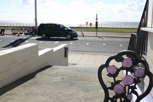 Een zwarte auto geparkeerd aan de kant van de weg, met uitzicht op de kust, een witte muur en een metalen hekwerk met paarse bloemen.