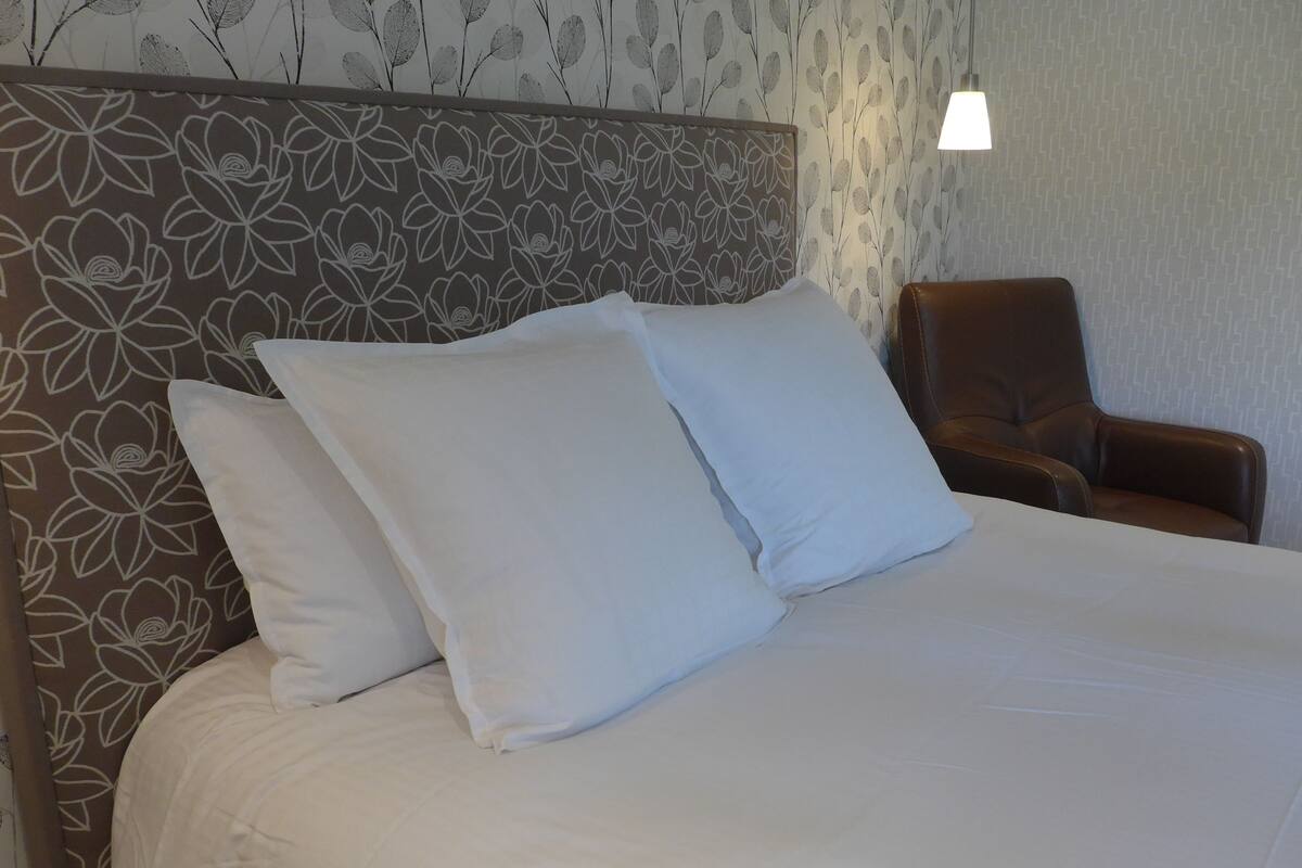 A neatly made bed with white linens and two pillows, a brown headboard with a floral pattern, and a brown leather chair to the right.