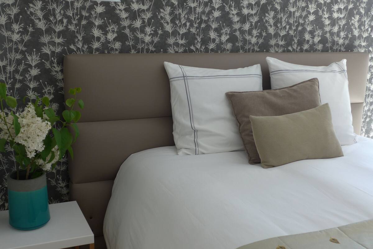 A neatly made bed with white linens and two pillows, a small bedside table with a vase of flowers, and a patterned wallpaper background.