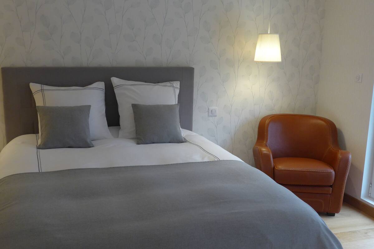 A neatly made bed with grey bedding and pillows, a brown leather armchair, and a wall with a leaf pattern.