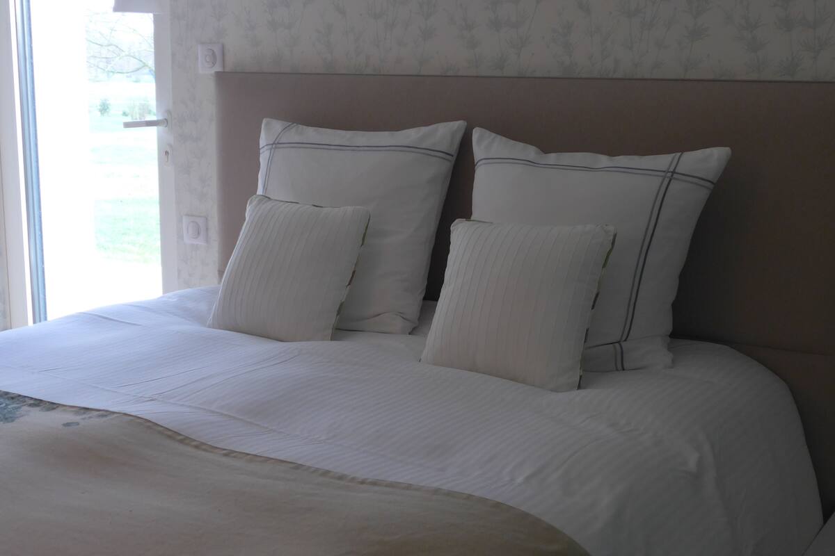A neatly made bed with white linens and two pillows, a headboard, and a window with a view of greenery.