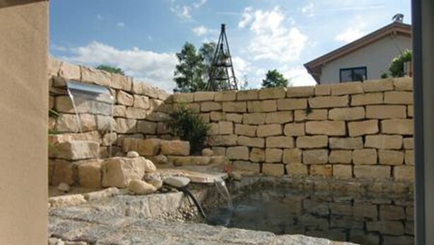 Un mur de pierre agrémenté d’un petit élément décoratif aquatique et, au fond, une structure métallique.