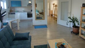 Un salon moderne avec un canapé, une table basse et un coin repas comprenant une table et des chaises. On aperçoit au fond une cuisine avec des meubles blancs et une cuisinière.