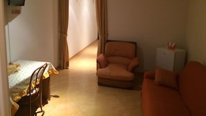 Een kleine, zwak verlichte kamer met een bed, een stoel, een bureau en een aan de muur bevestigde televisie.