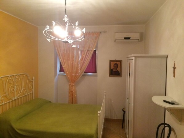 Un dormitorio con cama, ventana con cortinas, aire acondicionado instalado en la pared, una mesita de noche pequeña con un control remoto y una cruz colgada en la pared.