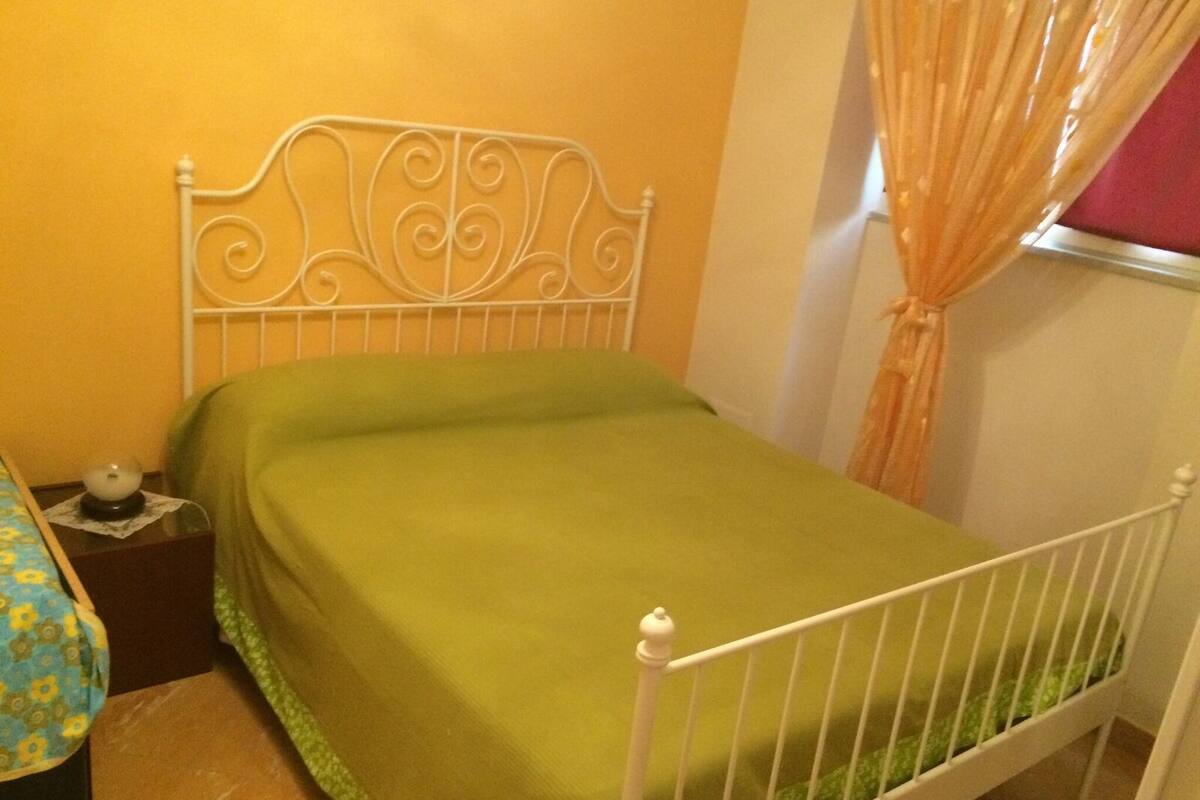 Een eenpersoonskamer met een metalen hoofdbord, een groene spreidekker en een nachtkastje met een lamp.