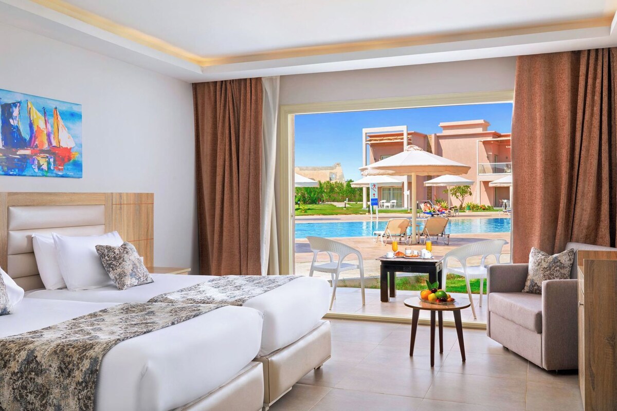 Habitación de hotel con cama, una mesa de desayuno y vistas a una piscina y un edificio.