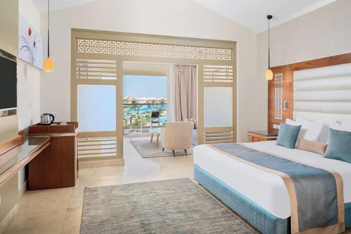 A hotel room with a large bed, a desk, a TV, and a view of a pool and resort buildings.