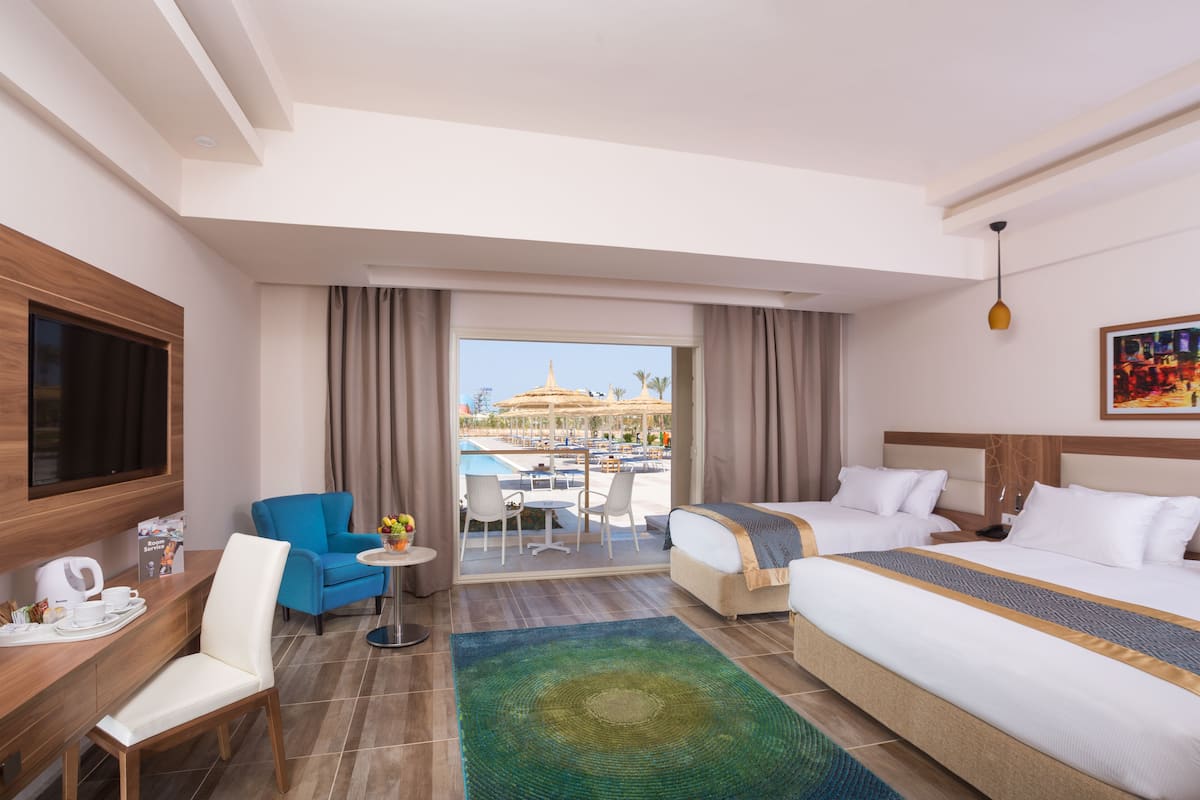 A hotel room with two beds, a TV, a desk, and a view of a pool and palm trees.