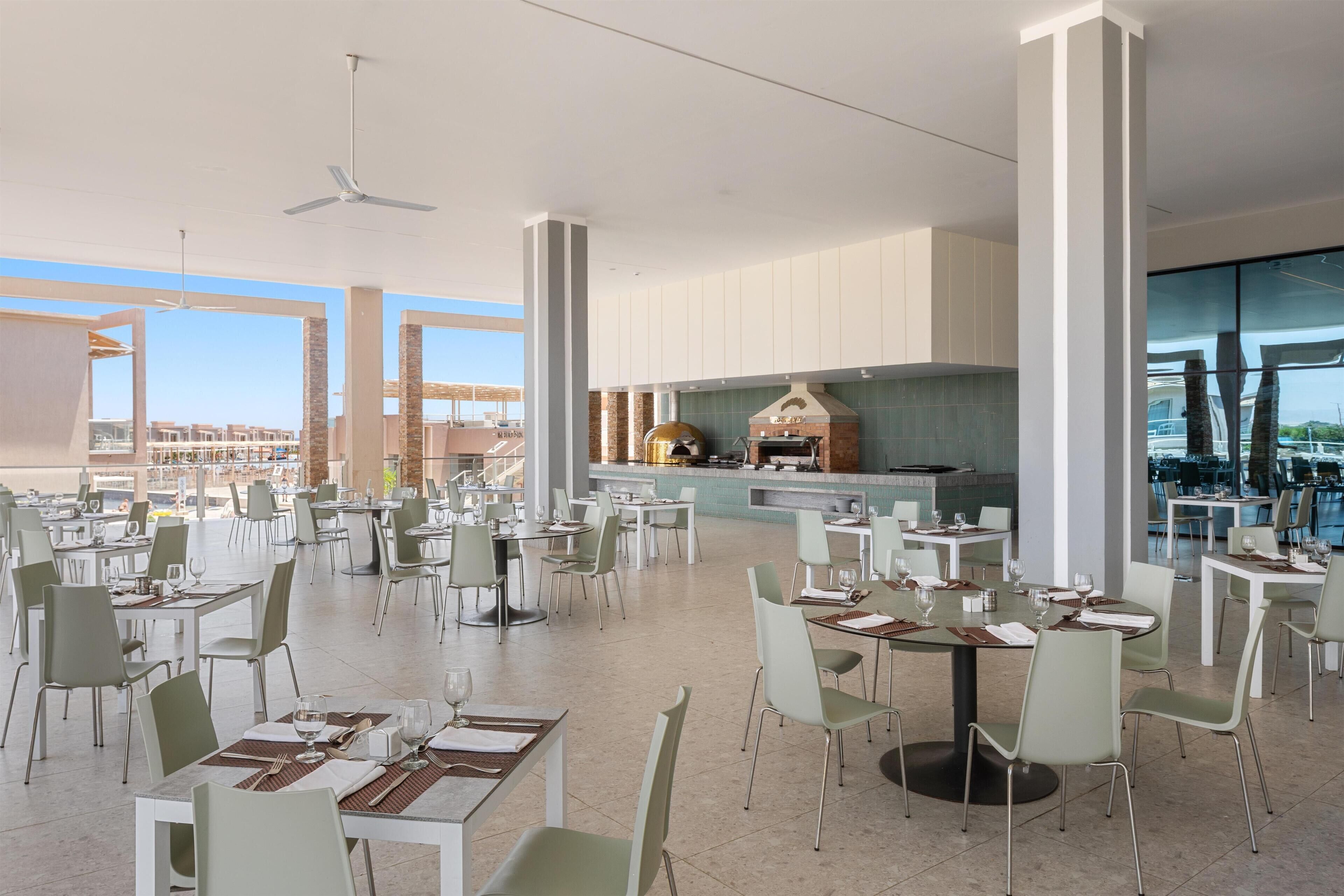 Un vaste espace de salle à manger avec de nombreuses tables et chaises, une grande cuisine équipée d’un barbecue et de grandes fenêtres offrant une vue sur la ville.
