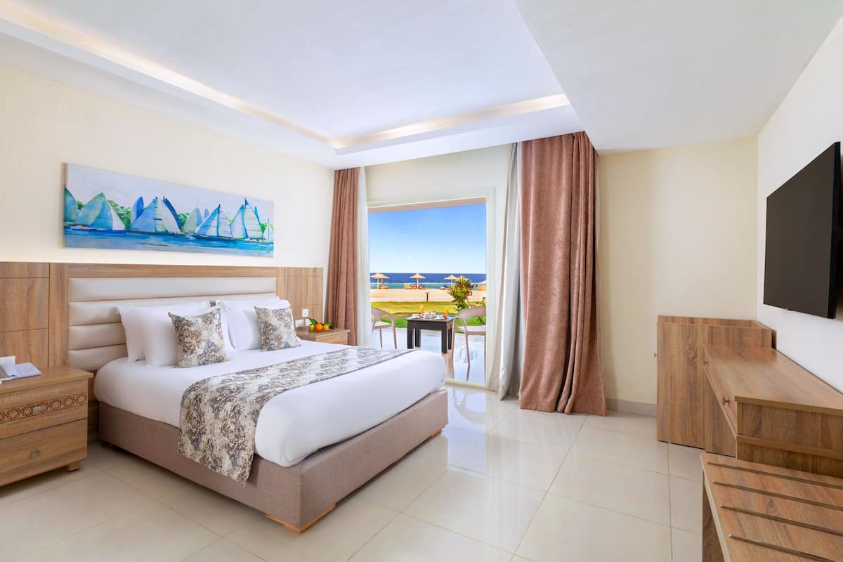 A hotel room with a bed, a desk, a TV, a painting of sailboats, and a view of the beach.