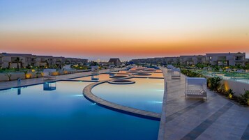 Zona de piscina en un resort con varias piscinas, sillas de descanso y un cielo al atardecer.