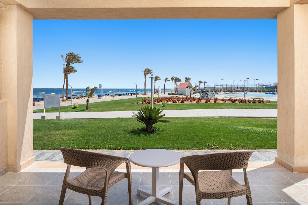 A balcony with wicker chairs and a small table, overlooking a well-maintained lawn with palm trees and a view of the beach and ocean.