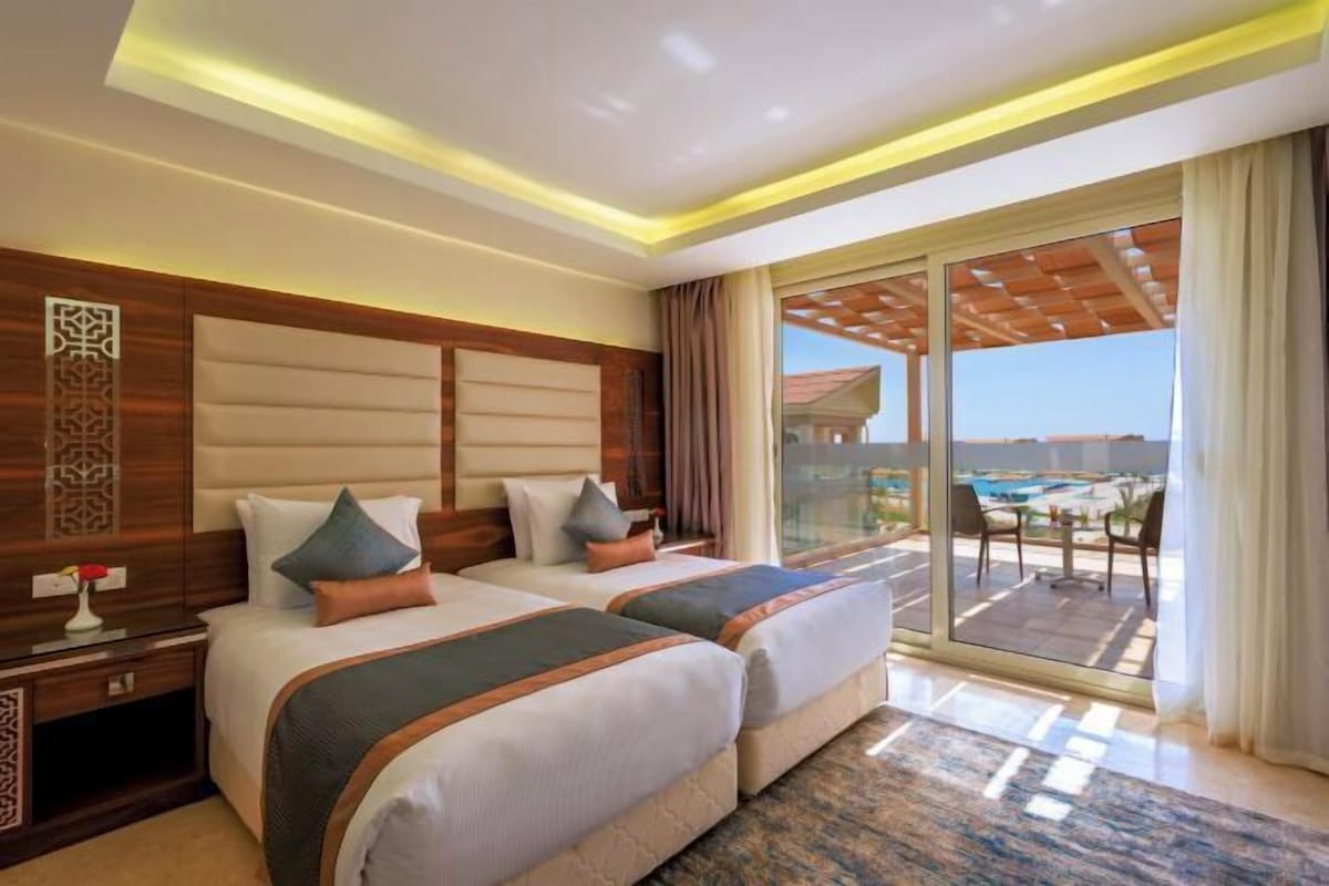 Habitación de hotel con dos camas, un ventanal con vista a la piscina y un balcón con mobiliario exterior.