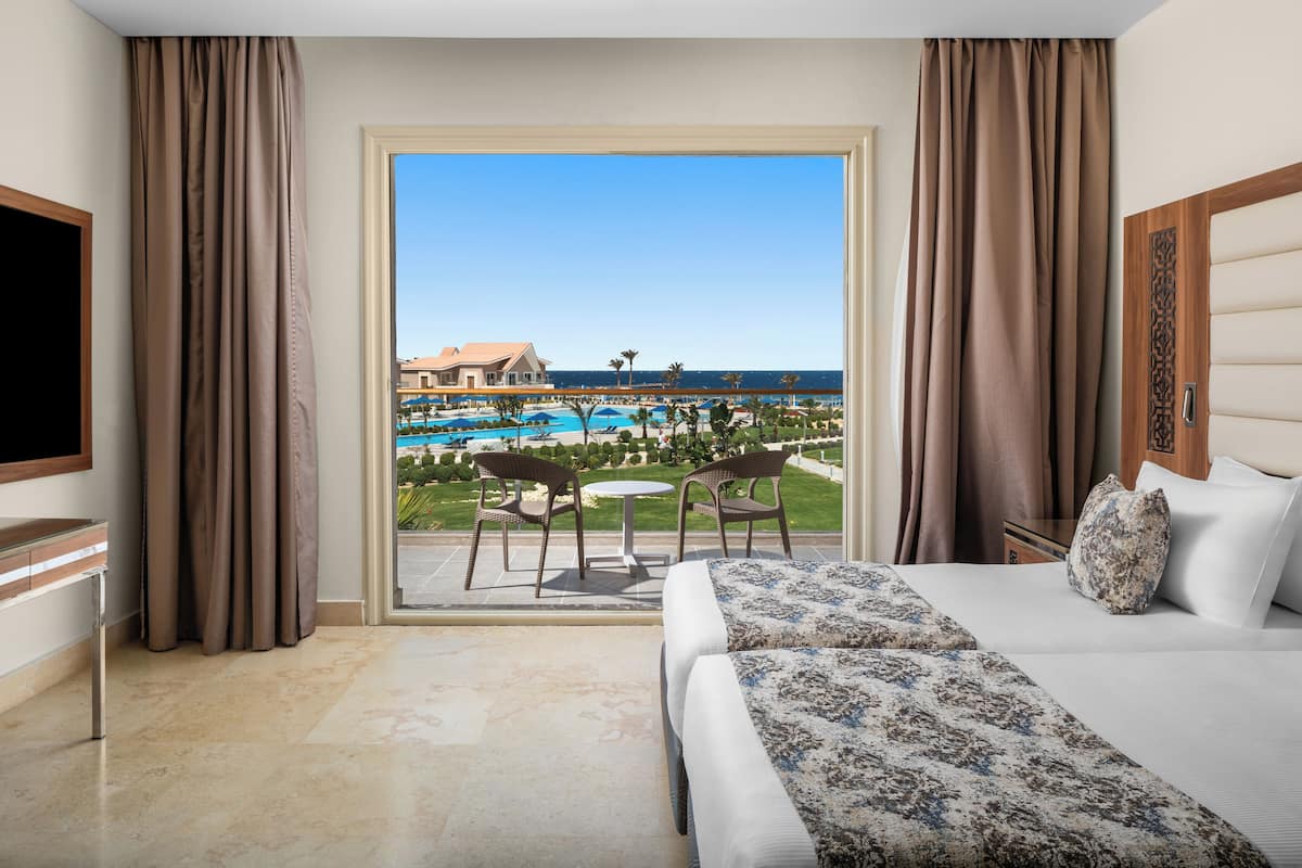 A hotel room with a bed, a TV, a desk, and a view of a pool and beach.