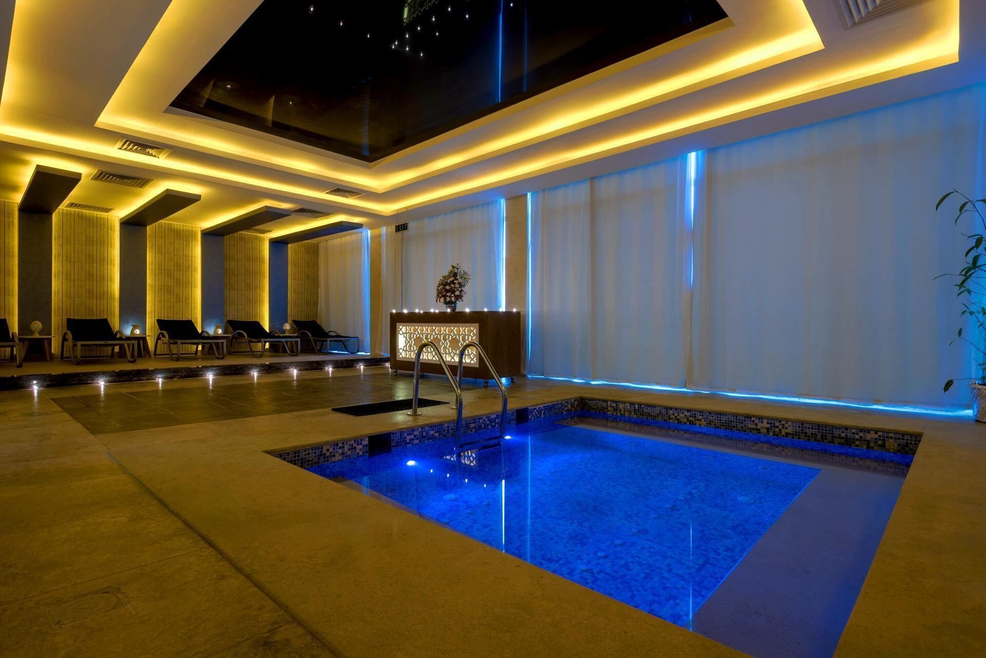 Piscina interior moderna con fuente de agua cristalina, rodeada de sillones de descanso y una zona de recepción.