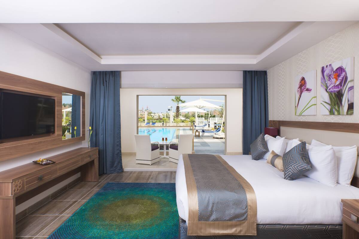 A hotel room with a large bed, a TV, a desk, and a view of a pool.