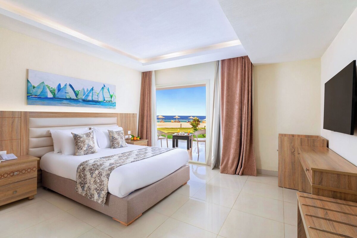 Habitación de hotel con cama, escritorio, televisor, un cuadro y vista a la playa.