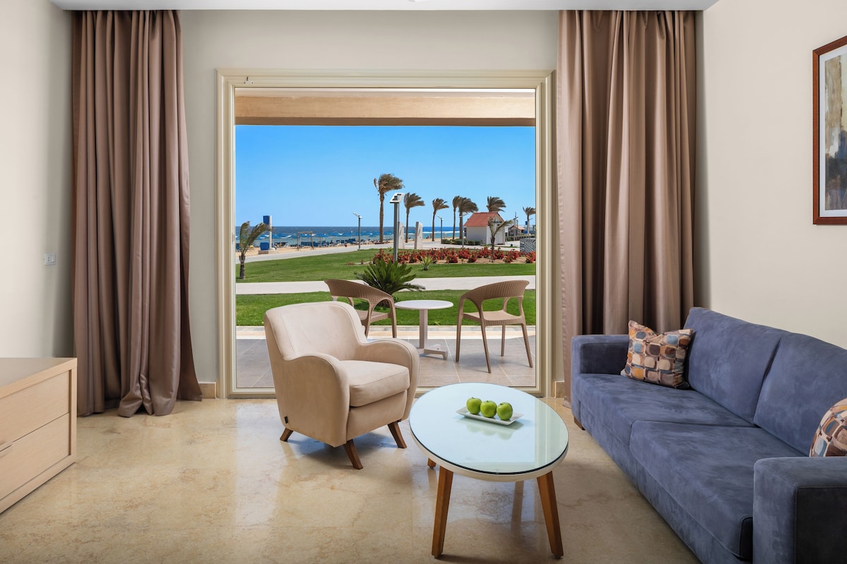Een kamer met uitzicht op het strand, een blauwe bank, een beige fauteuil en een glazen tafel met appels.