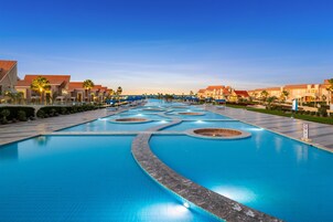 Una gran piscina de diseño moderno, rodeada de edificios tipo resort con techos rojos y palmeras.