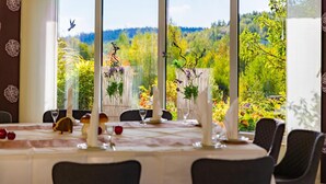 A dining area with a table set for four, large windows offering a view of a garden and trees. - Business Vital Hotel (Suhl)
