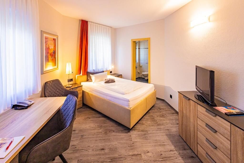 Habitación de hotel con cama, escritorio, silla, televisor y baño visible a través de una puerta abierta.