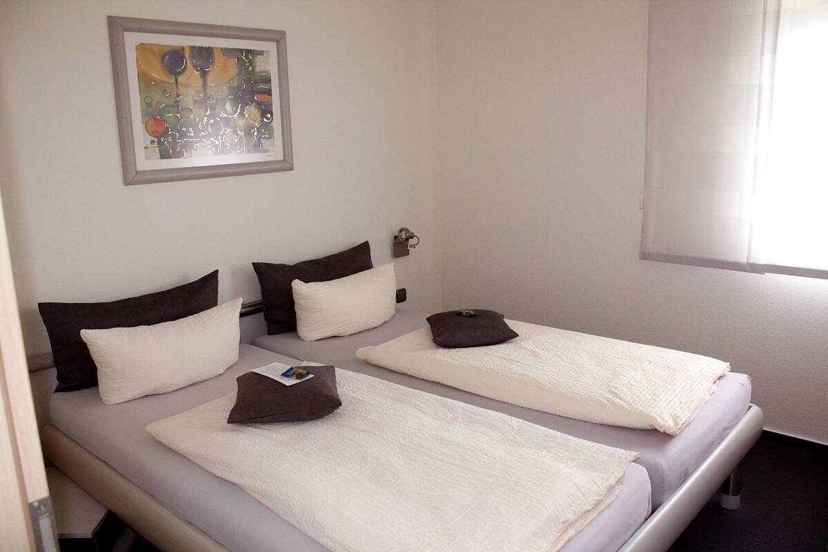 A hotel room with two single beds, each with a pillow and a blanket. There is a framed abstract painting on the wall above the beds.