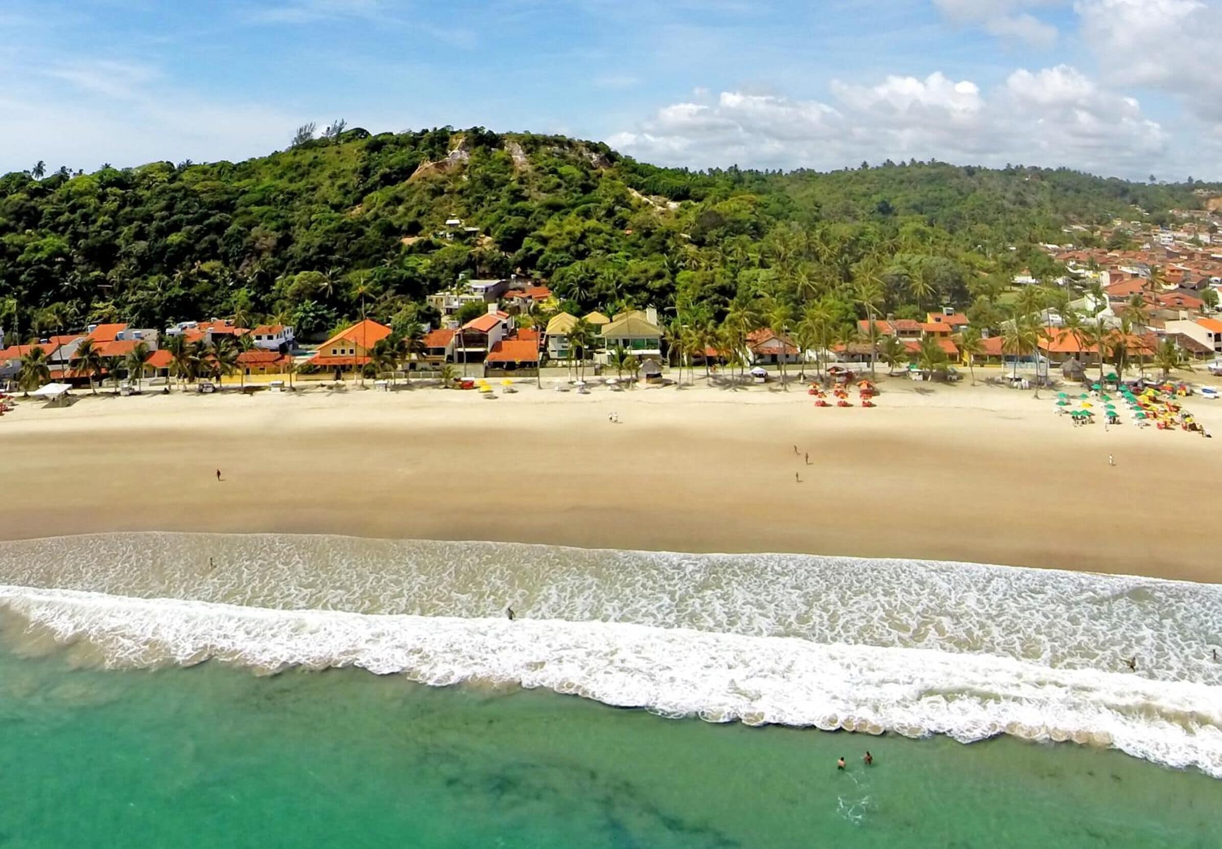 A beach with houses, clear turquoise water, and a hilly background.