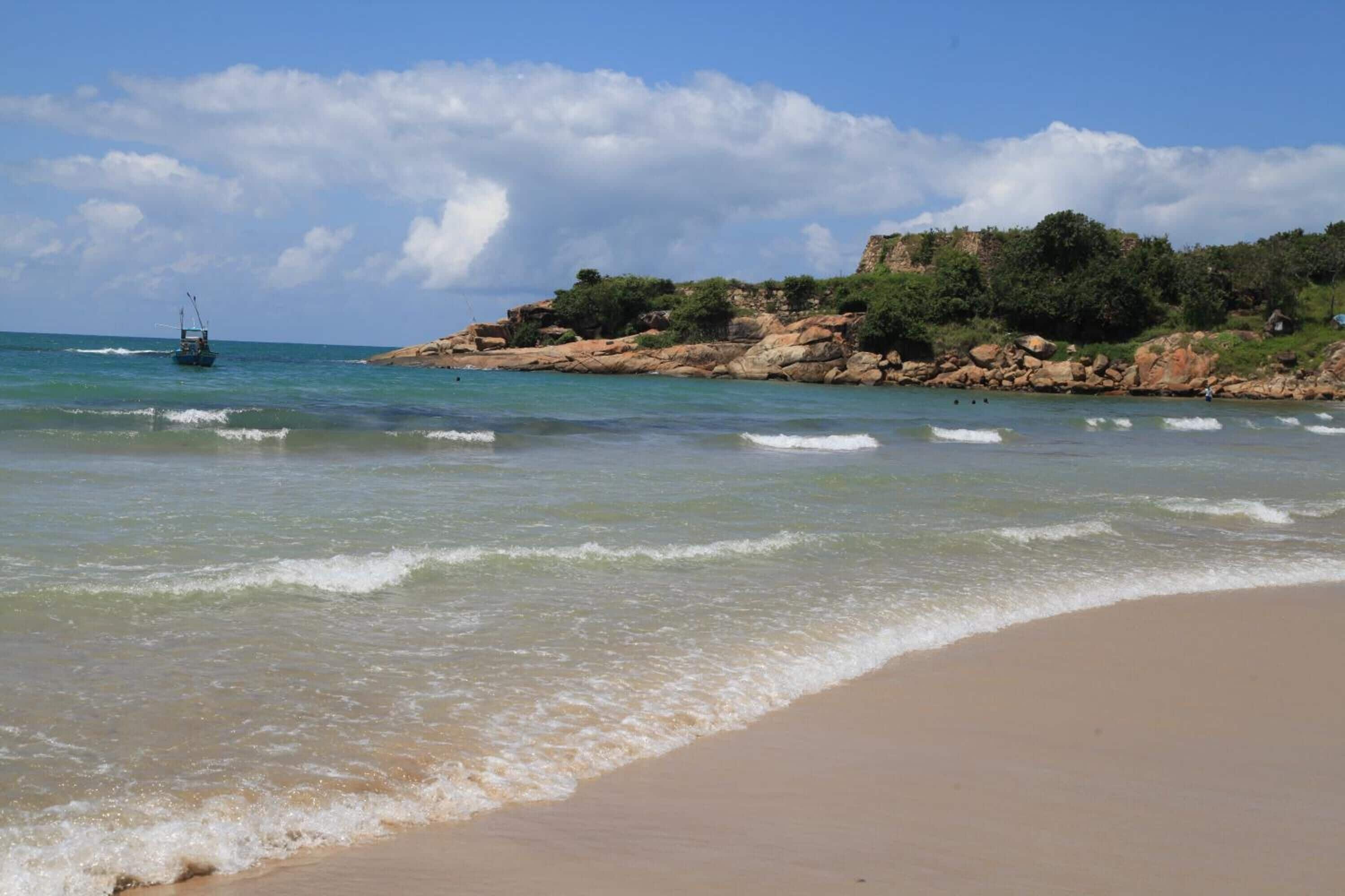 Una playa con aguas cristalinas, un afloramiento rocoso con vegetación y un barco a lo lejos.