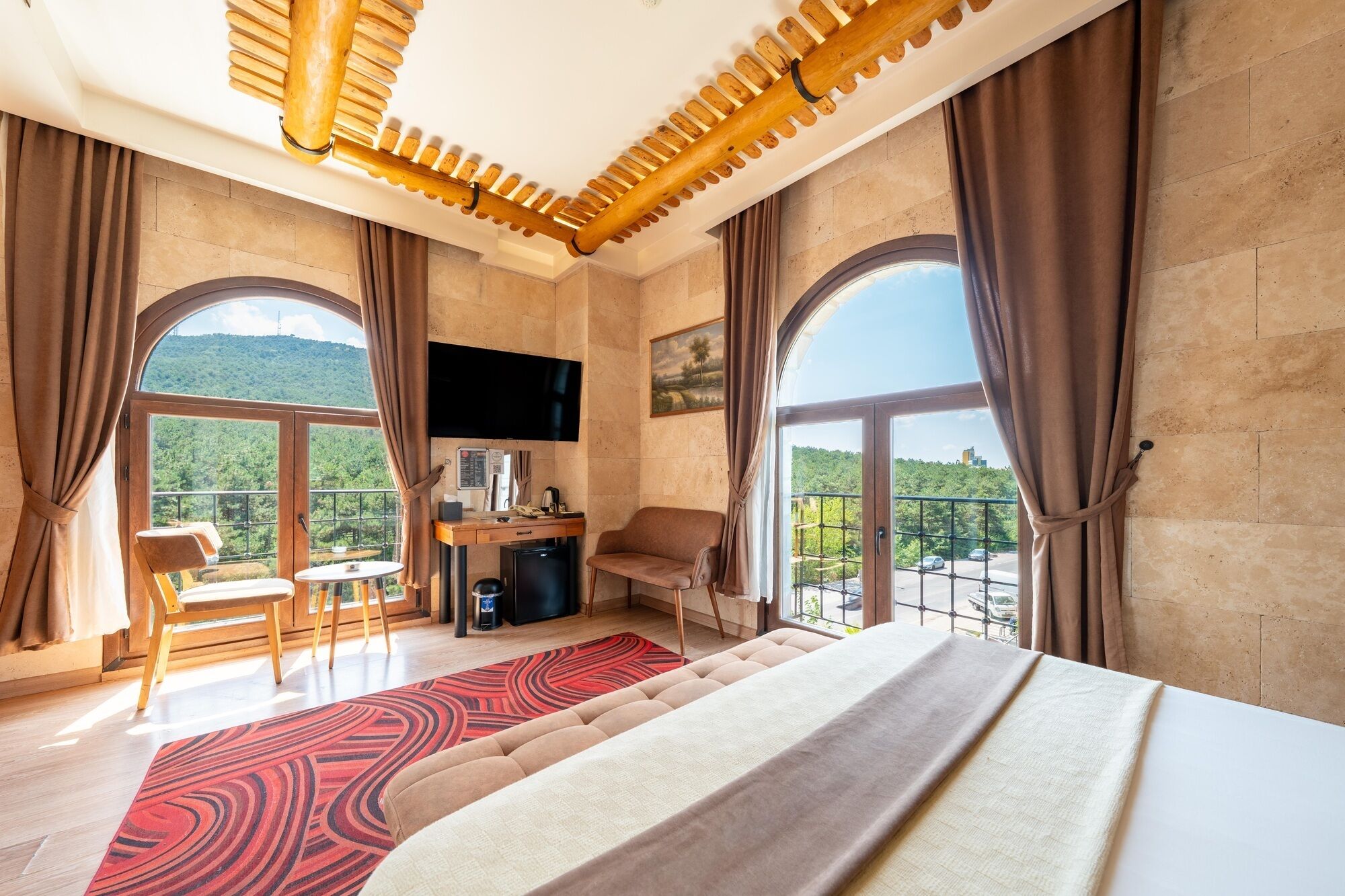 A hotel room with a bed, a desk with a chair, a TV, and large windows offering a view of the outdoors.