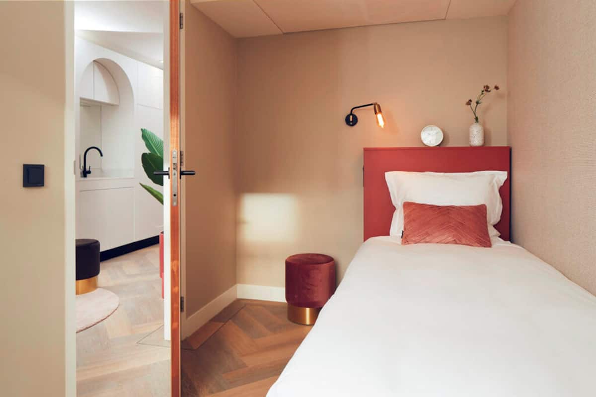 A bedroom with a bed, a bedside lamp, a small red stool, and a plant on the nightstand.