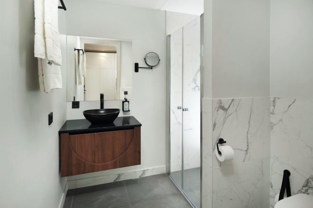 Un baño moderno con un lavabo negro, tocador de madera, espejo, cabina de ducha con mampara de cristal y paredes de mármol.