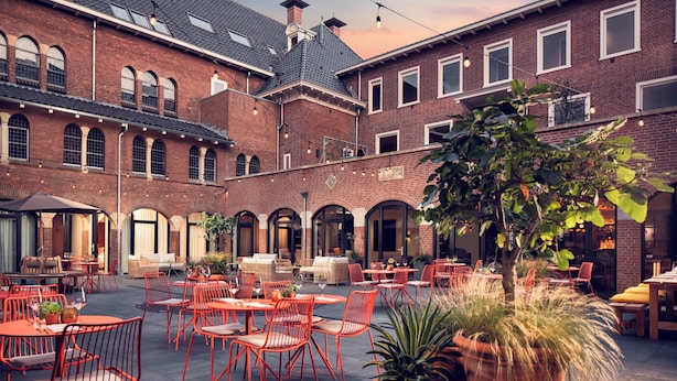 An outdoor courtyard with red metal chairs and tables, surrounded by brick buildings with arched windows and a large potted plant in the center. - The Anthony Hotel (Utrecht)