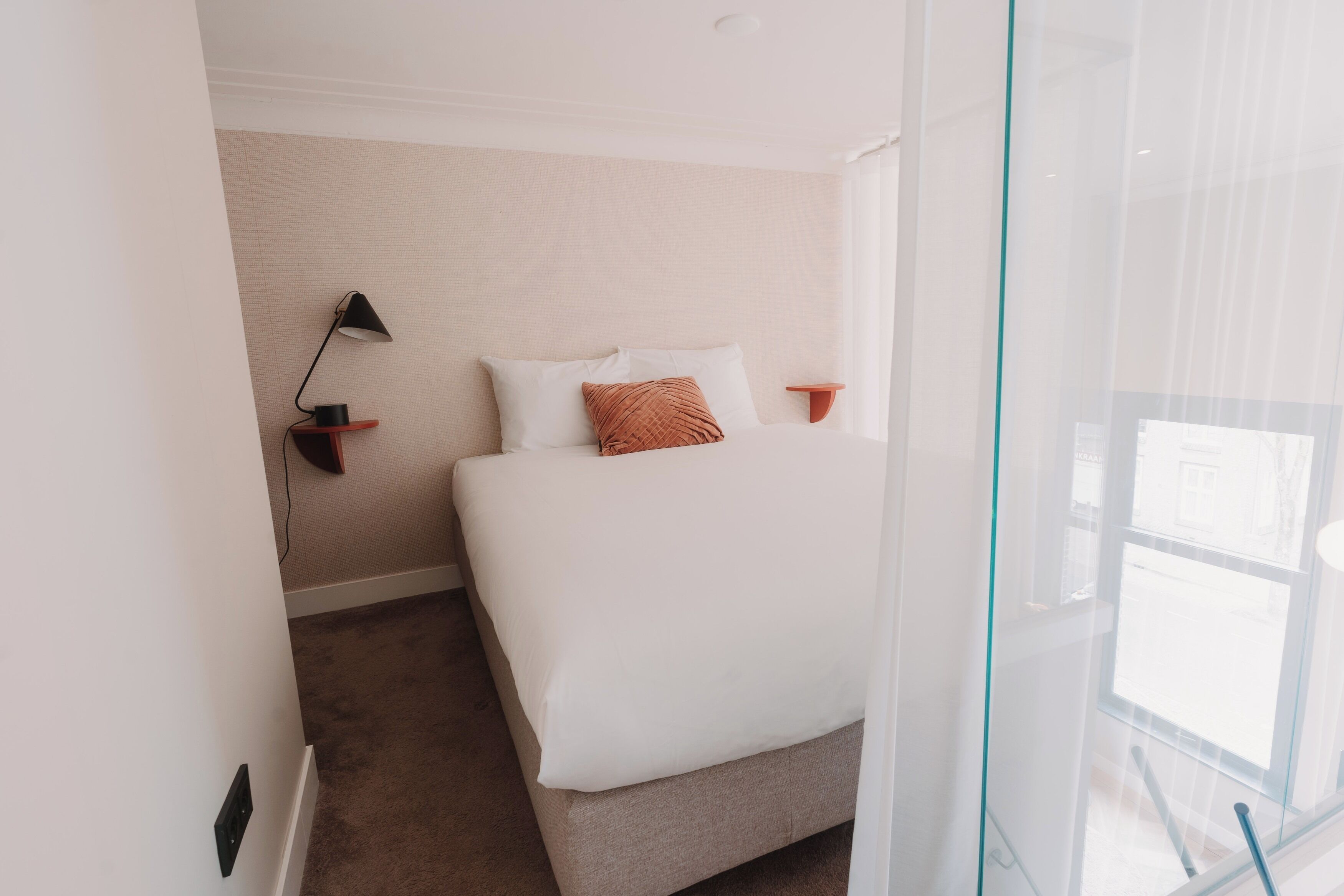 A hotel room with a bed, a bedside table with a lamp, a small shelf, and a window with curtains.