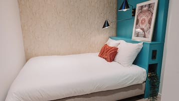 Ein Einzelbett mit weißer Bettwäsche, ein Türkis-Nachttisch mit einem gerahmten Bild und eine schwarze Stehlampe.