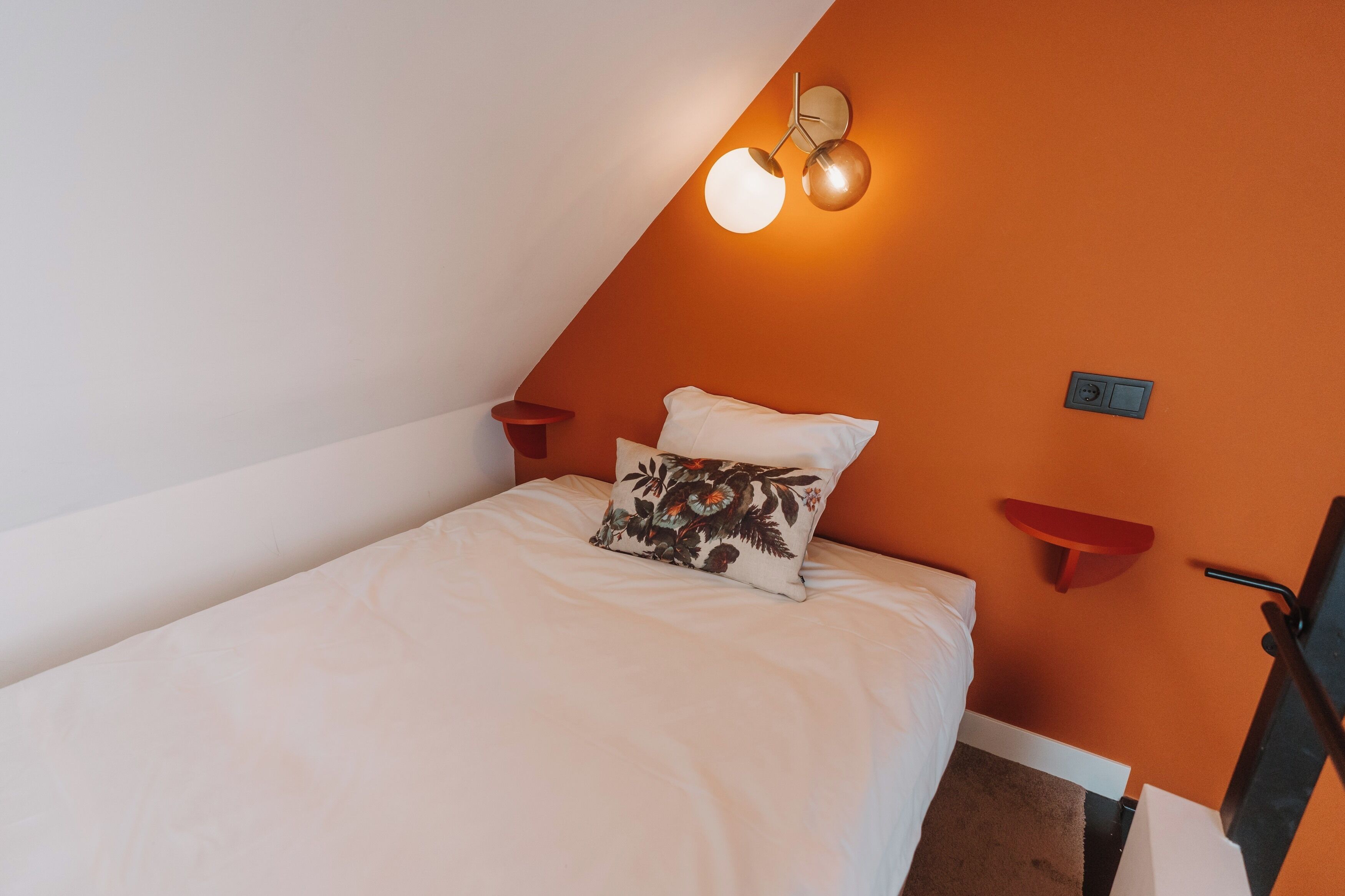 A bedroom with a white bed, a floral pillow, a red wall, and a wall-mounted light fixture.