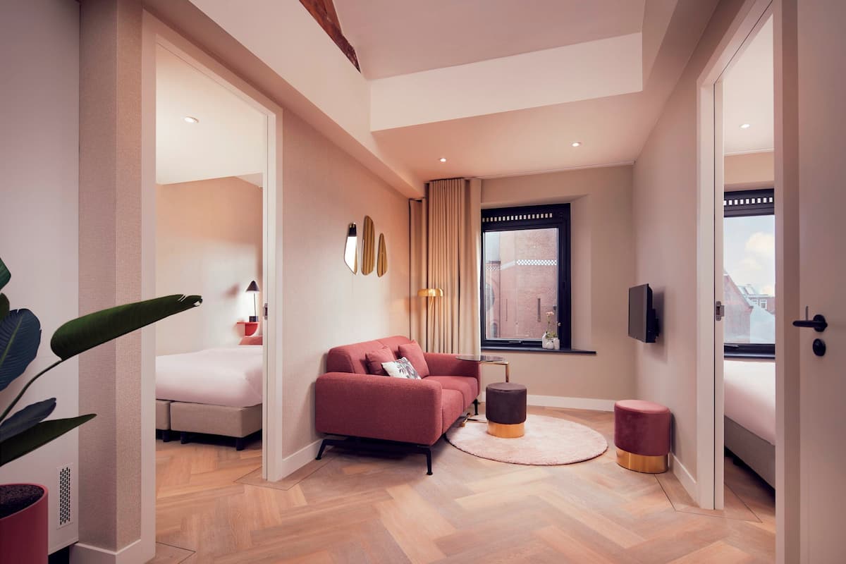 A modern hotel room with a sofa, a small round table, a potted plant, and a view of the city through a window.