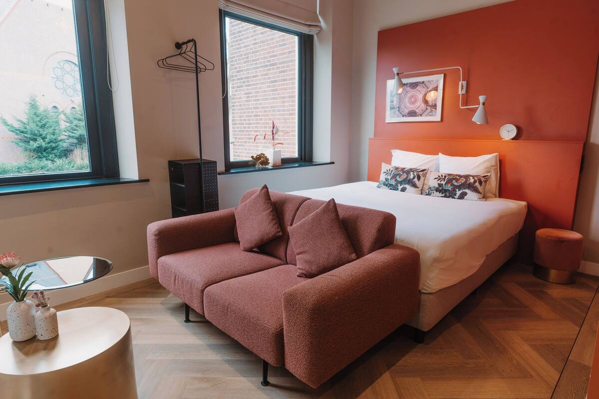 A modern bedroom with a large bed, a red sofa, a round side table, and a window with a view of a brick building.