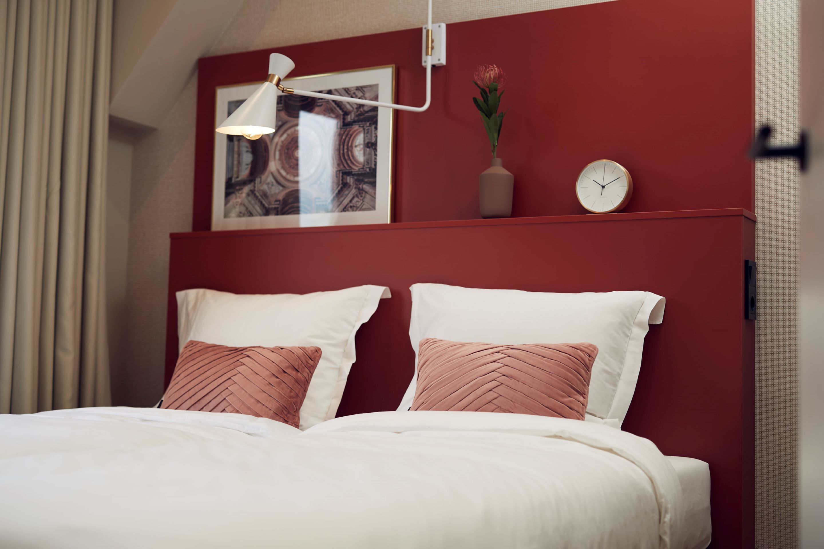 A bedroom with a red headboard, a framed picture, a clock, and a vase with flowers.