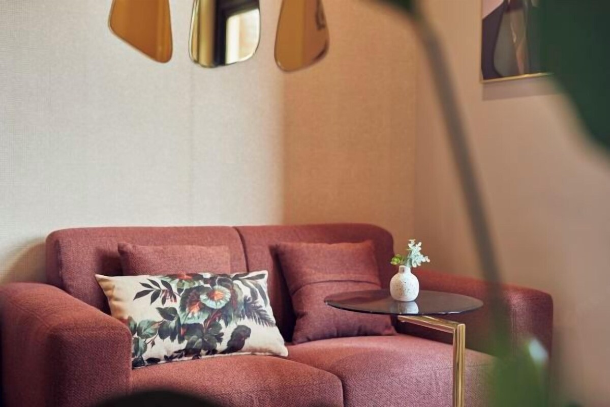 Un sofá rojo con un cojín floral, una mesita redonda con un florero y un espejo fijado a la pared.