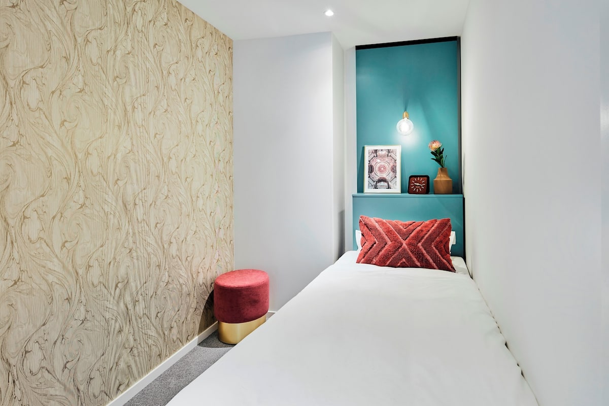 A bedroom with a white bed, a red ottoman, a teal accent wall, and a small shelf with decorative items.